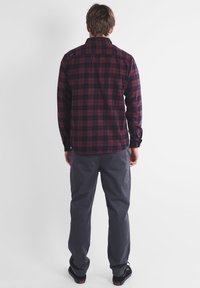 Chemise en flanelle à carreaux rouges et marine avec des manches longues, des poignets boutonnés et un dos droit. Associée à un pantalon gris et des chaussures noires.