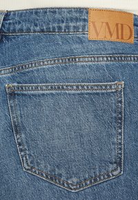 Denim jeans featuring a tan leather logo patch labeled "VMD," five-pocket design, and blue color with a faded texture.