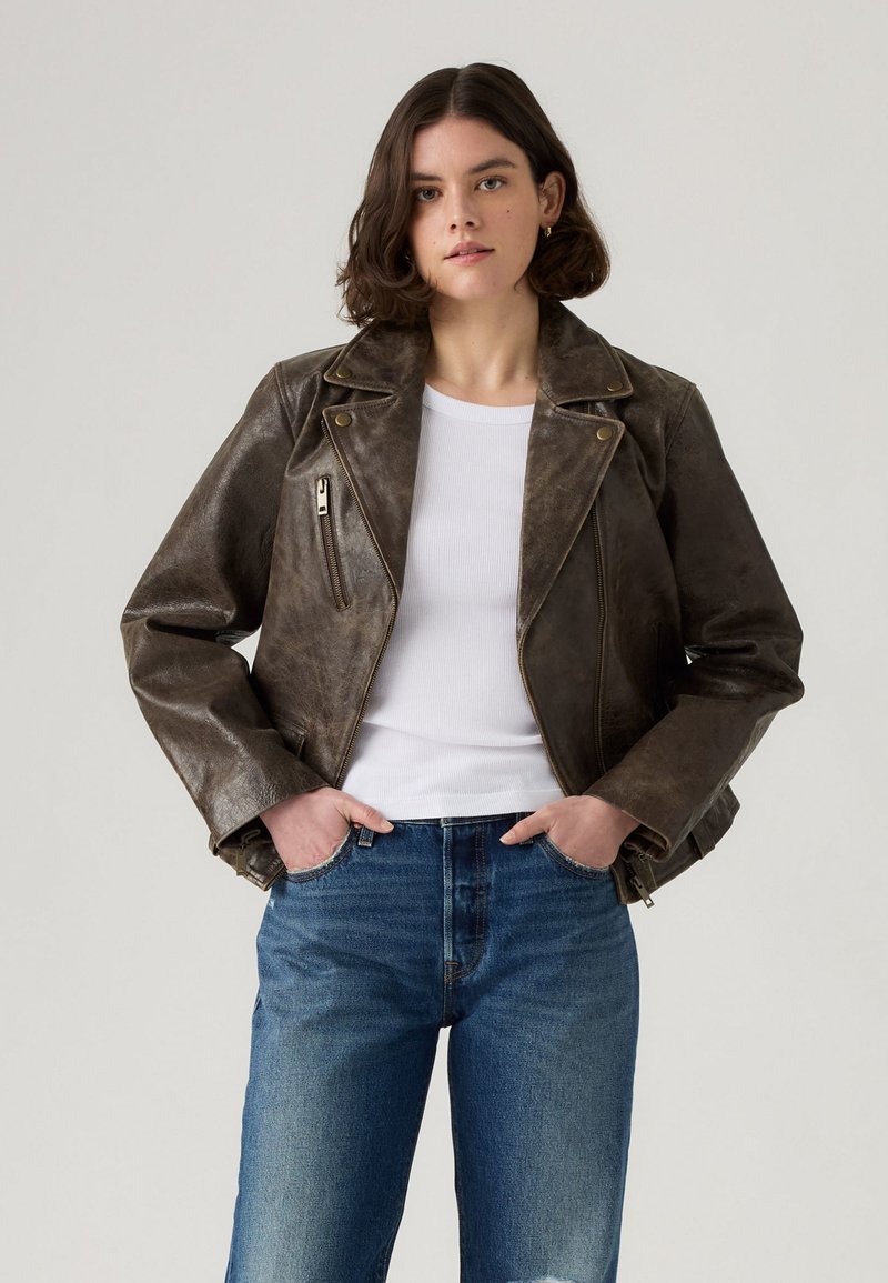 Veste en cuir marron de motard avec une finition légèrement usée, poche zippée et col à revers, associée à une chemise blanche et un jean bleu.