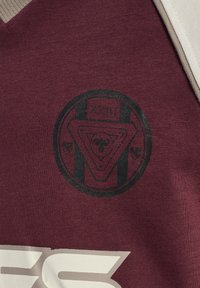 Vinrött tyg med en svart cirkulär logotyp med geometriska mönster och en dödskallemotiv, tillsammans med ljusbeige accenter och fet text nedanför.