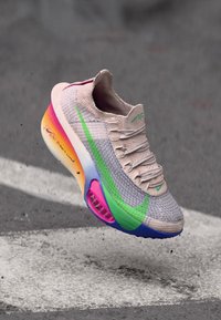 Lys grå sneaker med strikket overdel, grønne og rosa aksenter, og gradient såle. Har et strømlinjeformet design og snøreløs konstruksjon.