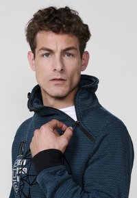 Chaqueta azul con estampado, con cremallera y cuello alto, puños acanalados y diseño de texto gráfico en la parte frontal. Se aprecia el detalle de la cremallera.