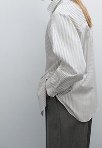 Persona in una camicia a righe chiare con bottoni e pantaloni scuri in piedi di lato con le mani nelle tasche su uno sfondo grigio.