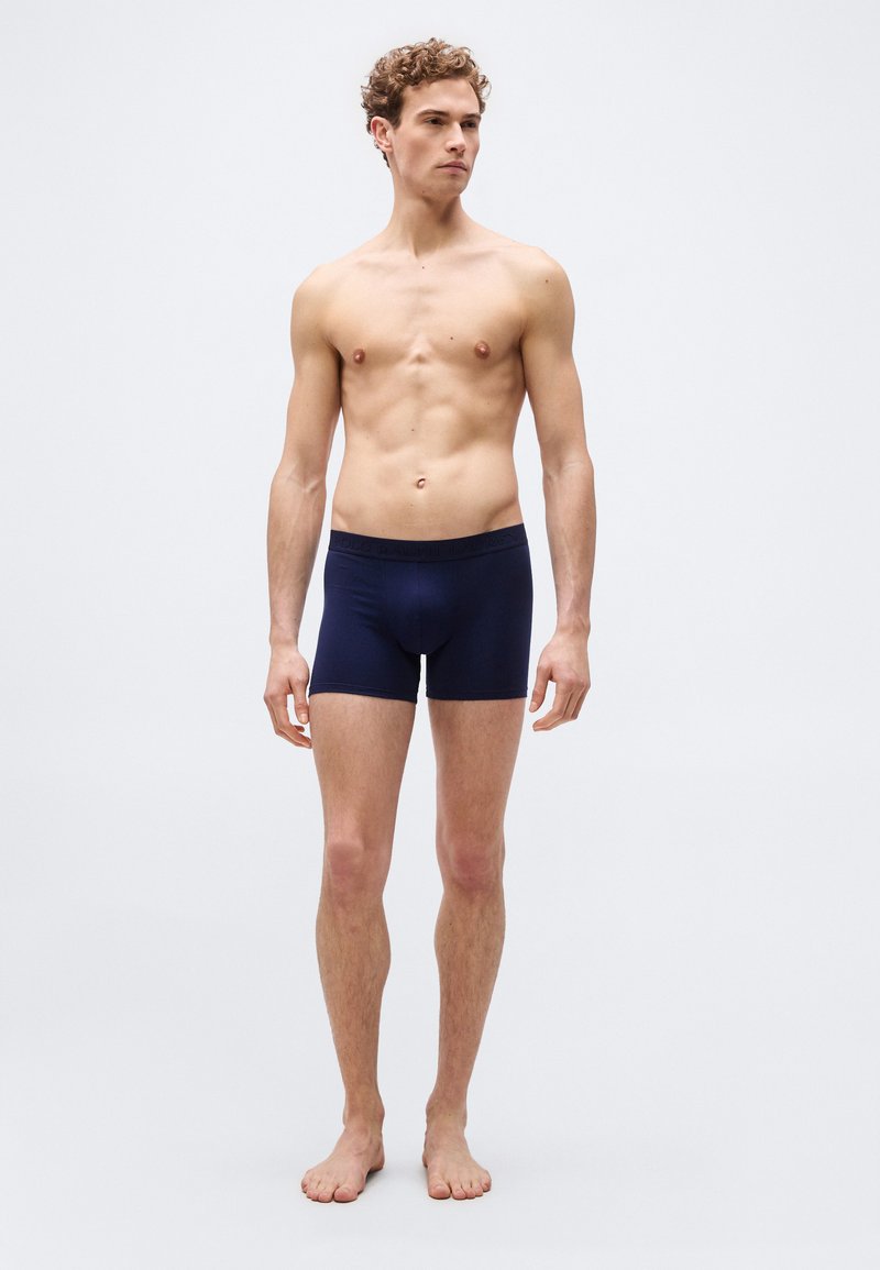 Νεαρός άντρας με σγουρά μαλλιά στέκεται ξυπόλυτος φορώντας σκουρόχρωμα ναυτικά μπόξερ, μπροστά σε απλό φωτεινό φόντο.