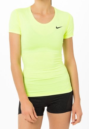 Vrouw draagt een nauwsluitend neongeel Nike T-shirt met korte mouwen en zwarte sportshorts, staand met ontspannen handen langs haar zij.