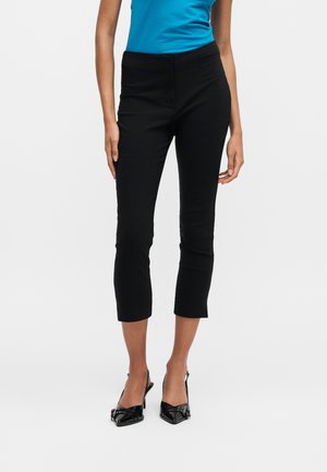 Kobieta nosząca dopasowane czarne krótkie spodnie, niebieski bezrękawnik i czarne czółenka z szpicem i klamrą, stoi na białym tle.