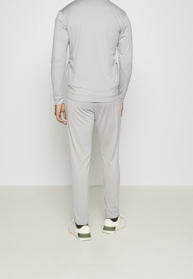 Homme de dos portant un survêtement gris clair et des baskets blanches avec un détail vert, debout devant un fond blanc uni.