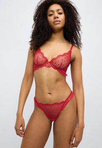 Σετ κόκκινης δαντέλας με διαφανές ύφασμα, κυματιστά άκρα και σχέδια καρδιών. Περιλαμβάνει σουτιέν με μπανέλα και τόνγκ που ταιριάζει.