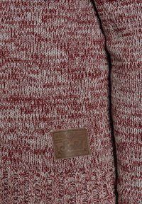 Jersey de punto rojo y gris con un patrón texturizado y una etiqueta de cuero marrón con "Solid" grabado en la esquina inferior derecha.