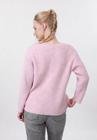 Femme aux cheveux blonds tressés portant un pull rose clair duveteux et un jean gris, debout avec une main sur la hanche, de dos.