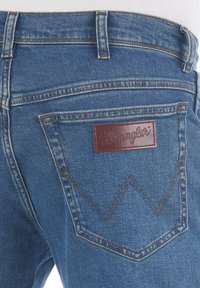 Jeans in denim blu con un'etichetta in pelle, caratterizzati da una particolare cucitura a zigzag sulla tasca posteriore e un design classico a gamba dritta.