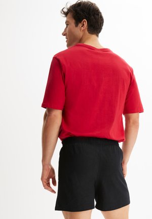 Rotes Baumwoll-T-Shirt mit kurzen Ärmeln, kombiniert mit schwarzen Shorts mit elastischem Bund. Der Stoff hat eine weiche Textur und eine lockere Passform.