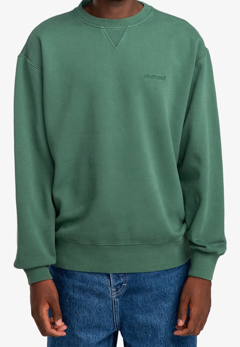 Personne portant un sweat-shirt vert col rond avec le logo "element" sur la poitrine et un jean bleu, debout devant un fond blanc.
