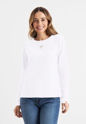 Young woman smiling wearing a white long-sleeve shirt with a small heart detail and blue jeans against a plain background.