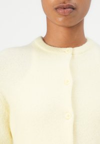 Cardigan giallo lavorato a maglia con scollo rotondo, texture morbida e tre bottoni sul davanti. Il colore è pallido, aggiungendo un calore sottile.