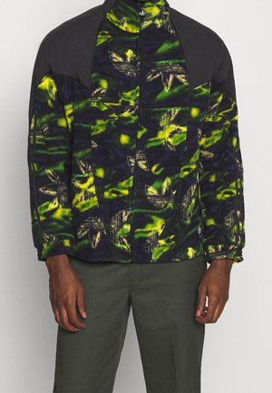 Man wearing a black and neon green patterned fleece jacket with dark green pants, standing against a plain background.