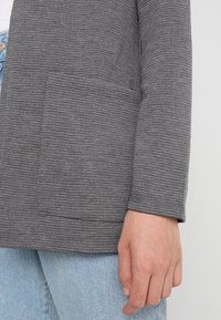 Szary kardigan z teksturowanym prążkowanym wzorem, otwarty z przodu, z jedną naszytą kieszenią po lewej stronie. Casualowy krój, w połączeniu z jasnymi dżinsami.