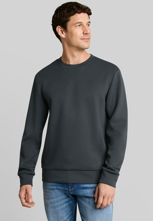 Uomo che indossa una felpa a maniche lunghe grigio scuro semplice e jeans blu, che guarda leggermente verso destra su uno sfondo grigio chiaro.