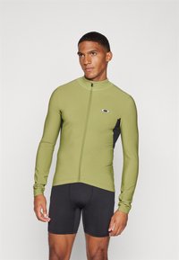 Haut de sport à manches longues vert olive avec fermeture éclair sur le devant, panneaux latéraux noirs et logo. Associé à un short noir ajusté. Tissu lisse et extensible.