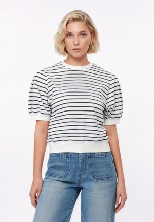 Blondynka w białym swetrze z czarnymi poziomymi paskami i bufkami na rękawach, w połączeniu z wysokim stanem niebieskich dżinsów, stojąca na białym tle.