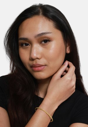 Bracciale d'oro con un design a catena attorcigliata indossato al polso; il modello ha i capelli lunghi e scuri e indossa un top nero.