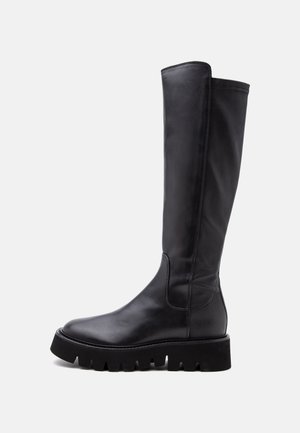 Botte haute en cuir noir avec semelle épaisse et rainurée et bout arrondi, vue de profil sur un fond uni clair.