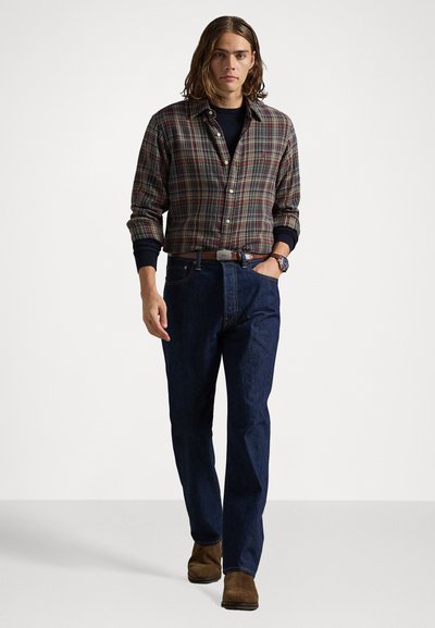 Geruite flanellen shirt in groene en rode tinten, gecombineerd met donkerblauwe jeans en bruine suède enkellaarsjes. Bevat een horloge om de pols.