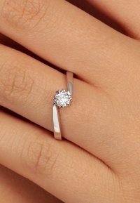 Silberring mit einem dünnen Band, ausgestattet mit einem runden, funkelnden Diamanten, der in einem blumenartigen Krappen-Design gefasst ist. Texturierte Haut im Hintergrund.