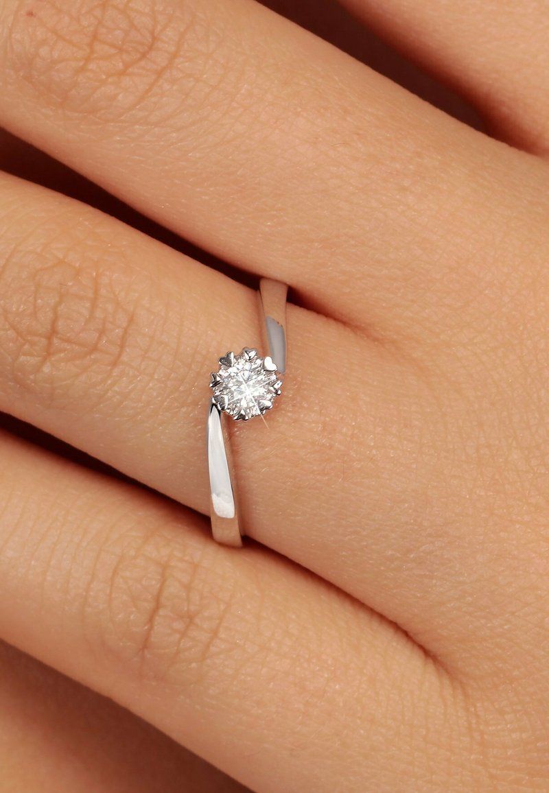 Silberring mit einem dünnen Band, ausgestattet mit einem runden, funkelnden Diamanten, der in einem blumenartigen Krappen-Design gefasst ist. Texturierte Haut im Hintergrund.