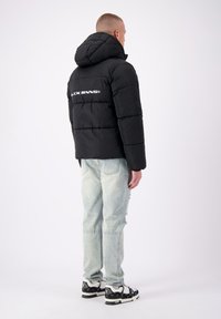 Zwarte pufferjack met een capuchon, met gestikte delen en een wit logo op de achterkant. Gecombineerd met lichtblauwe versleten jeans en gebloemde sneakers.