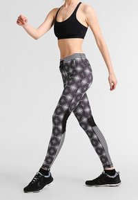 Reggiseno sportivo nero e leggings a fantasia con un design a reticolo di punti bianchi. Accentuati da pannelli in rete e abbinati a scarpe da ginnastica nere.