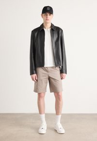 Sort læderjakke med lynlås over en hvid crew-neck trøje, kombineret med beige shorts og hvide sneakers. Tilbehøret inkluderer en sort kasket.
