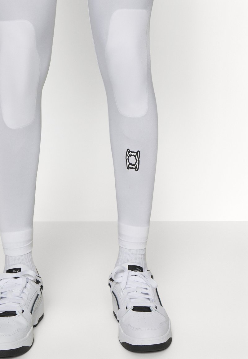 Leggings bianchi con imbottitura alle ginocchia e un logo sulla gamba inferiore, abbinati a sneakers bianche e nere e calzini bianchi. Texture liscia.