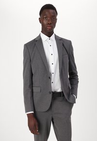 Costume gris avec veste et pantalon assortis, tissu texturé, fermeture à un bouton, chemise blanche avec boutons foncés, poche sur la veste.