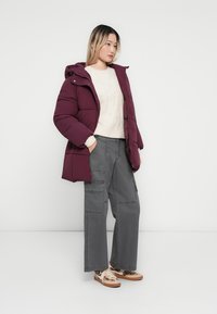 Vinröd puffjacka med huva, med ett kviltat mönster. Bärs över en krämfärgad tröja och grå cargobyxor. Ljusfärgade sneakers fulländar outfiten.
