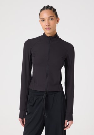 Vrouw met ingevlochten haar die een nauwsluitend zwart jack met ritssluiting en losse zwarte broek met trekkoord draagt, tegen een effen witte achtergrond.