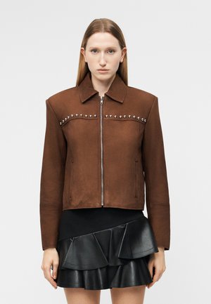 Γυναίκα που φοράει καφέ τζάκετ με φερμουάρ και ασημένιες καρφίτσες στο στήθος και μαύρη δερμάτινη φούστα με στρώσεις και βολάν.