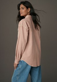 Cămașă roz deschis cu mânecă lungă, cu o croială lejeră, detalii cu manșete cu nasturi și un tiv curbat, combinată cu blugi din denim albastru, foarte lejeri.