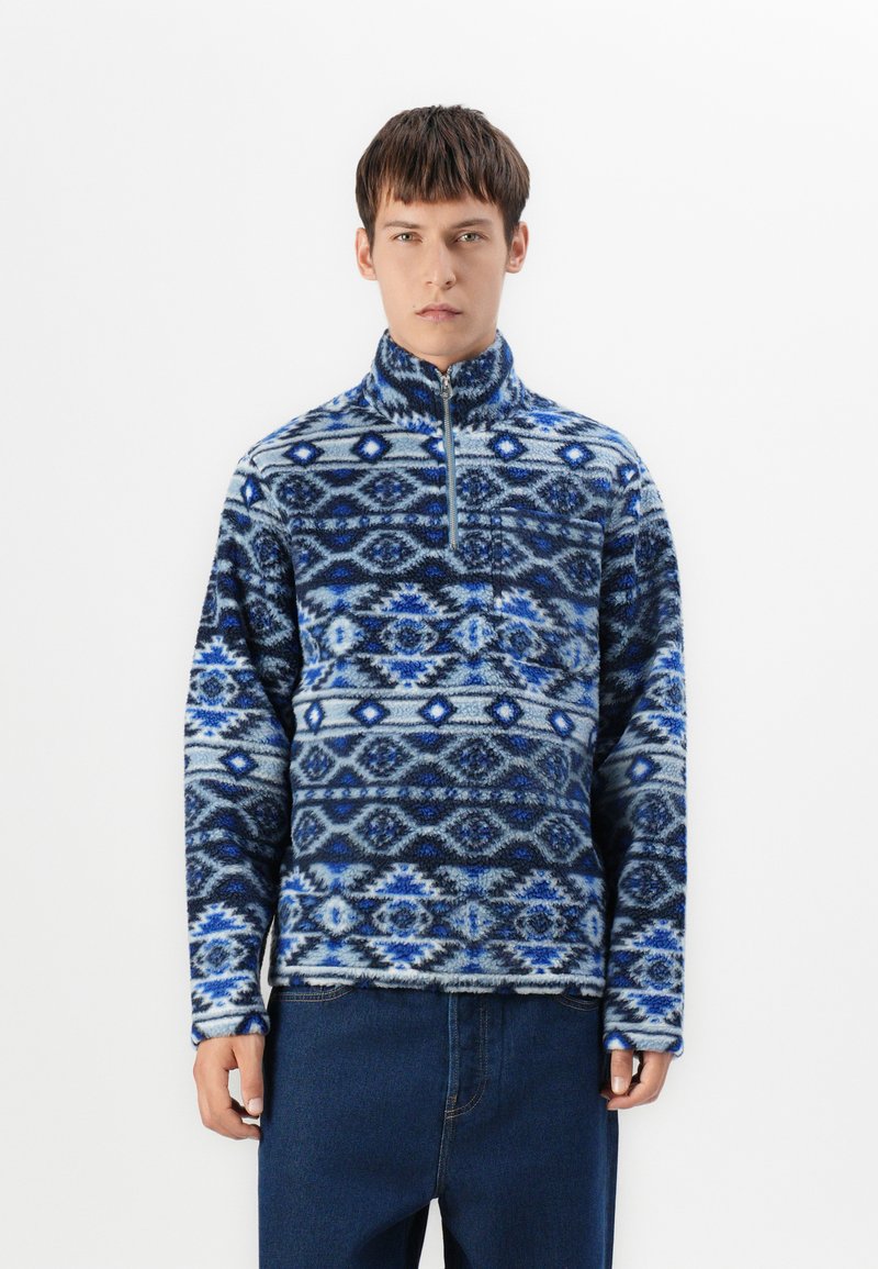 Fleece-pullover med et geometrisk mønster i blå og grå nuancer, med lynlås i halsen, lange ærmer og en blød, struktureret overflade.