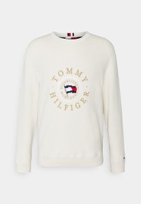 Weißes Sweatshirt mit langen Ärmeln, ausgestattet mit einem goldenen runden Logo, das den Schriftzug "TOMMY HILFIGER" und ein farbiges Flaggenmotiv in der Mitte zeigt.