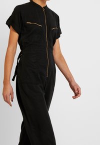 Zwarte jumpsuit met korte mouwen, twee voorzakken met rits, een centrale gouden rits en een cinched taille met strikken. Gladde stoftextuur.