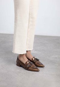 Zapatos de cuero marrón con punta puntiaguda, detalles texturizados y hebilla decorativa. Combinados con pantalones de color claro con dobladillo desflecado.