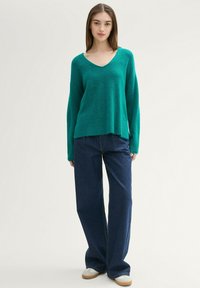 Grüner V-Ausschnitt-Pullover aus einem weichen, strukturierten Strick mit überschnittenen Schultern, kombiniert mit weit geschnittenen dunkelblauen Jeans und lässigen Schuhen.