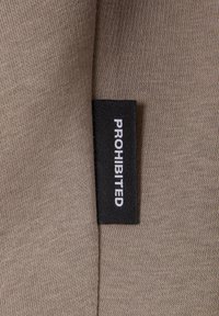 Beige szövet, sima textúrával, fekete címkével, ami fehér betűkkel azt írja: "TILTVA". A címke bele van varrva a szövetbe.