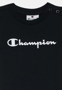 Schwarzes Baumwoll-T-Shirt mit Rundhalsausschnitt, das ein weißes "Champion"-Logo über der Brust und Druckknöpfe an der Schulter hat.