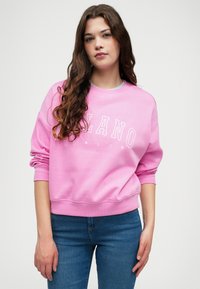 Sweatshirt rose à manches longues, col rond, avec une inscription graphique devant "MILANO ITALIA" en blanc. Associé à un jean bleu, texture lisse.