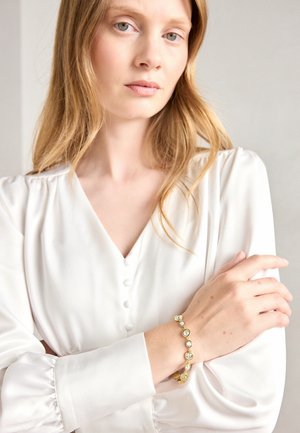 Gullarmbånd med runde klare edelstener, båret på en lys hudarm. Modellen har på seg en hvit bluse med V-hals og knappedetaljer.