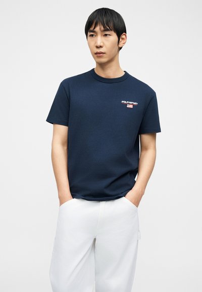 Άνδρας που φοράει μπλε σκούρο μπλουζάκι Polo Sport και άσπρο παντελόνι, στέκεται με τα χέρια στις τσέπες μπροστά σε απλό φόντο.