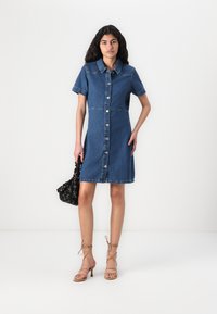 Vestido de camisa de ganga, mangas curtas, fecho de botões, cor azul clara, bolsos laterais, até ao joelho, combinado com uma bolsa preta e saltos com tiras.