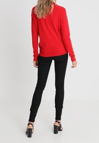 Rød langærmet sweater parret med sorte skinny jeans og sandaler med blokhæl. Sweateren har en glat tekstur og en afslappet pasform.